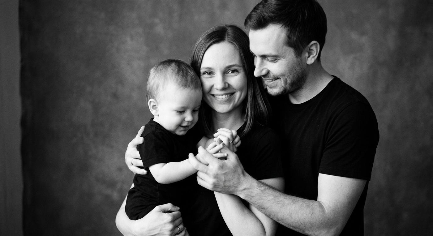 Создай hyper realistic black & white family portrait по референсу, сохранив лицо и черты внешности без изменений. Кинематографичное студийное фото семьи: мама держит малыша на руках, отец стоит рядом и нежно обнимает их обоих, прижимая ребёнка к себе. Мама смотрит прямо в камеру с мягким любящим взглядом, малыш играет руками родителей, отец смотрит на ребёнка с теплотой и заботой.

Все одеты в минималистичные чёрные футболки. У девушки длинные прямые волосы. Атмосфера уютная, эмоциональная и очень живая, акцент на семейную связь и естественные эмоции.

Мягкий студийный свет создаёт красивые тени и подчёркивает текстуру кожи, чёрно-белая обработка, мягко размытый фон, shallow depth of field. Cinematic photography, soft film grain, authentic emotions, realistic skin texture, ultra detailed, 8K, no AI look, no plastic skin, minimalist luxury style.